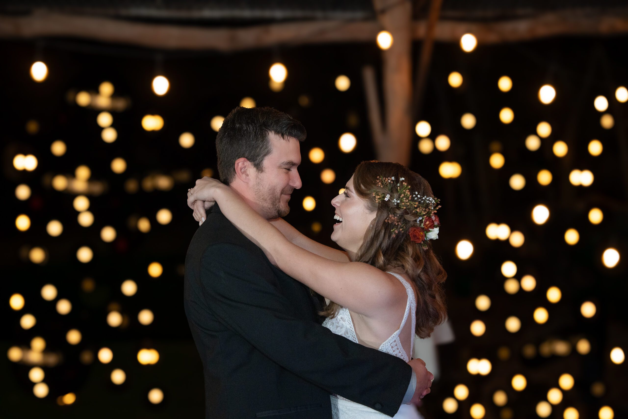 Bride and Groom dancing while looking at each other and smiling at night in front of twinkle lights