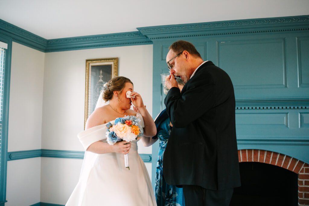 Father of the bride and bride stand facing each other and dabbing their eyes with tissues after first seeing one another in their wedding clothes.