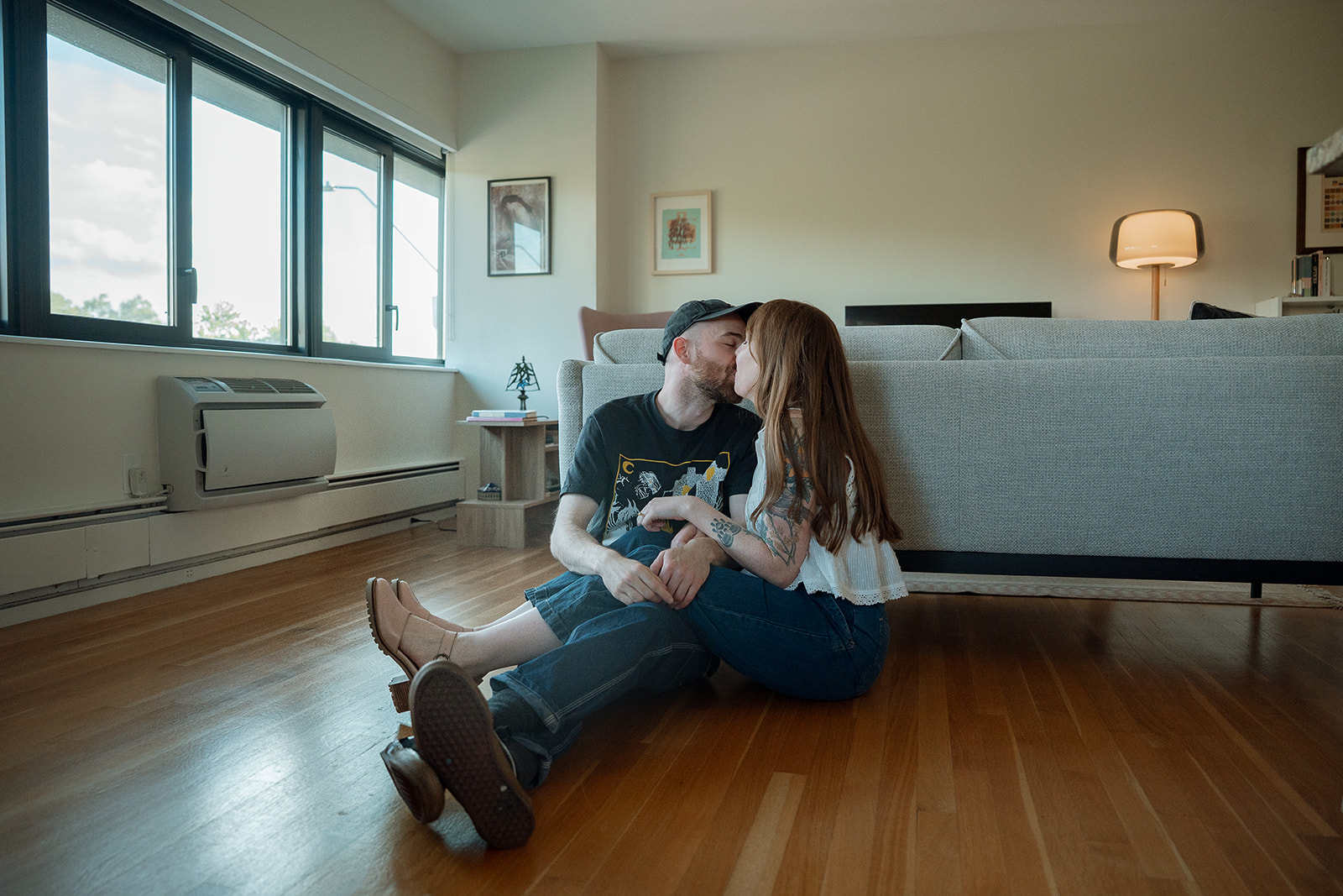A man and woman sit on the floor with their backs against a couch kissing