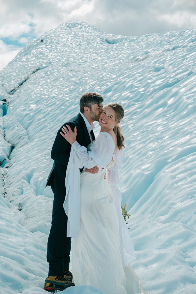 A man in a black suit kisses a woman in a white wedding dress atop a glacier in Alaska