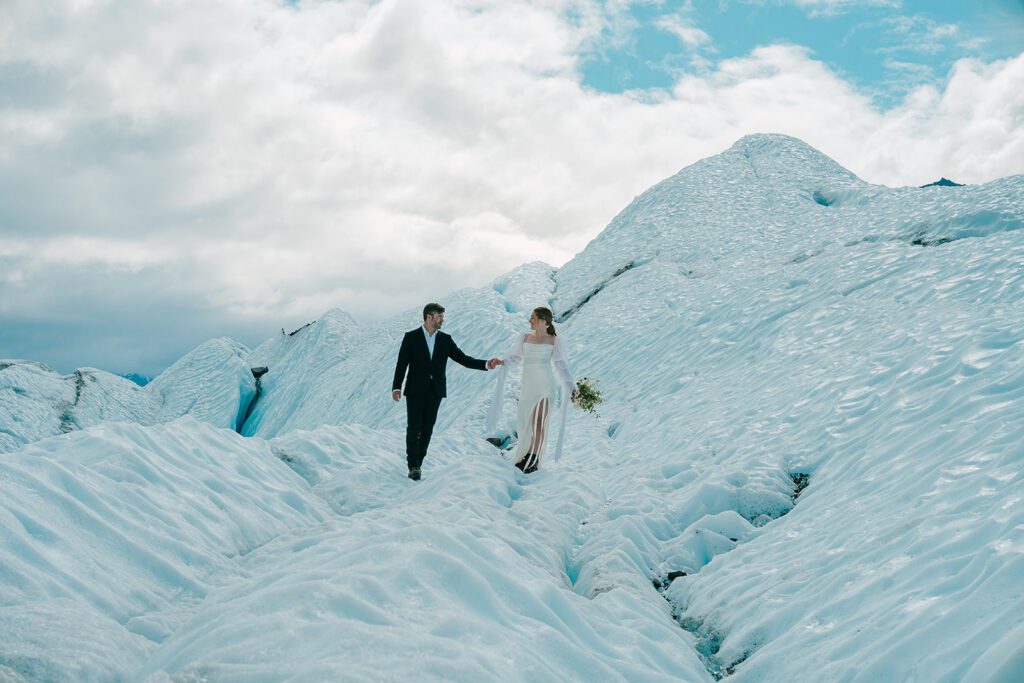 A man and woman in wedding clothing hold hands on top of a glacier in Alaska. The woman holds a bouquet and they look at each other. 