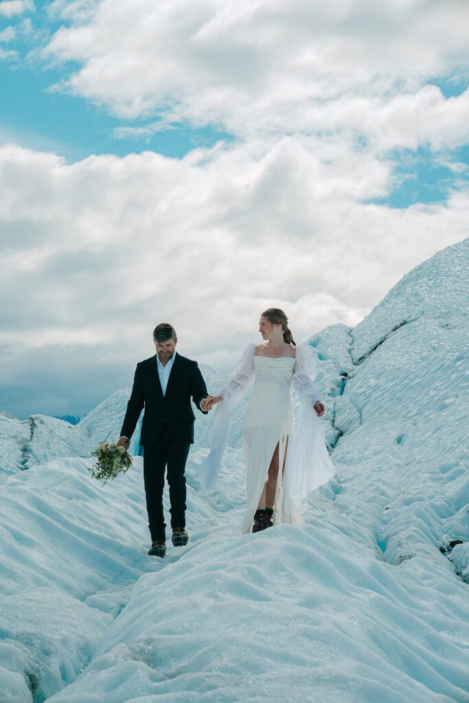 A man and woman in wedding clothing hold hands on top of a glacier in Alaska. The woman holds a bouquet and they look at the camera. 