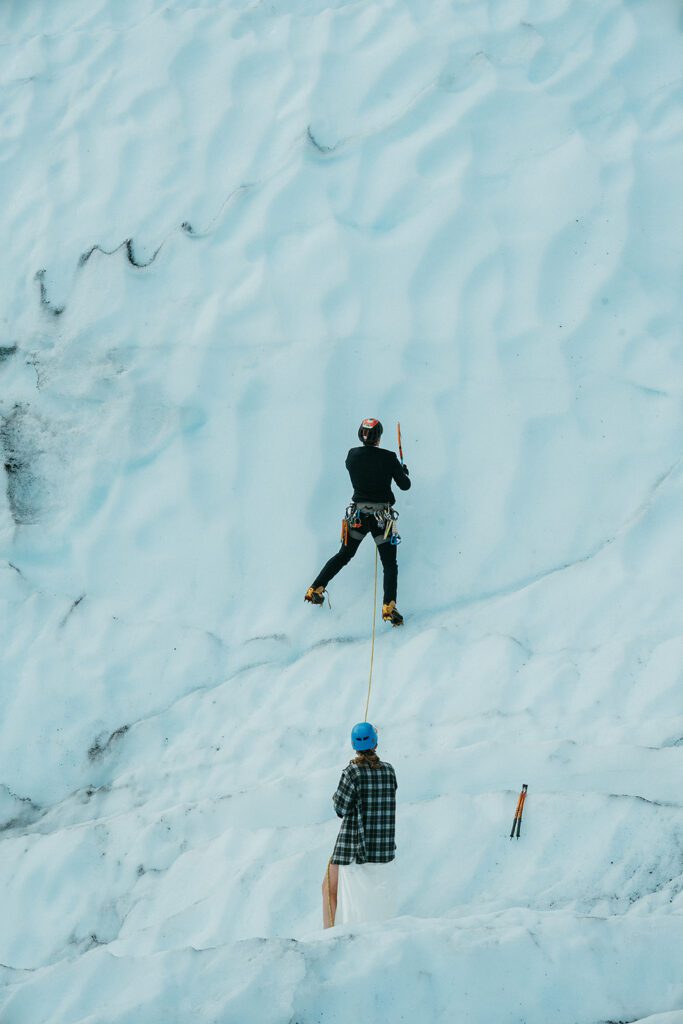 A man and woman in wedding clothes climb up the side of a Glacier in alaska. 