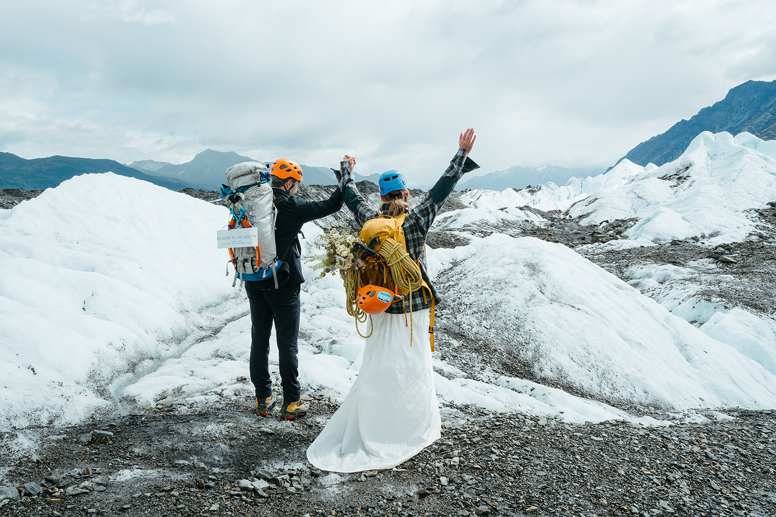 A man and woman in wedding clothes hold their hands in the air on top of Matanuska Glacier in Alaska. They are wearing backpacks with ice climbing gear inside and out.