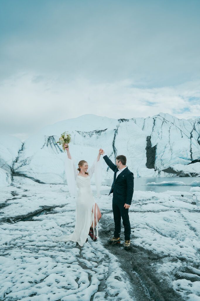 A man and woman in wedding clothing cheer and raise their hands on a glacier in Alaska. 