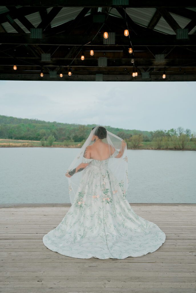 A woman in a floral wedding gown stands in front of a lake and holds her veil to the sides