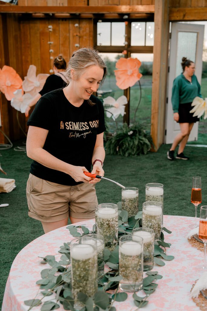 A woman stands next to a round table and lights candles placed in glass containers.