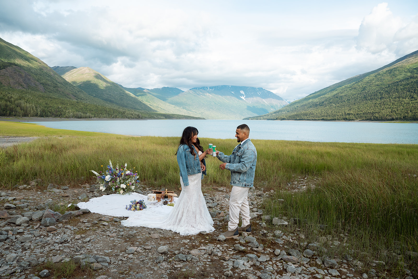A bride and groom stand outside in front of a lake with mountains in the distance.