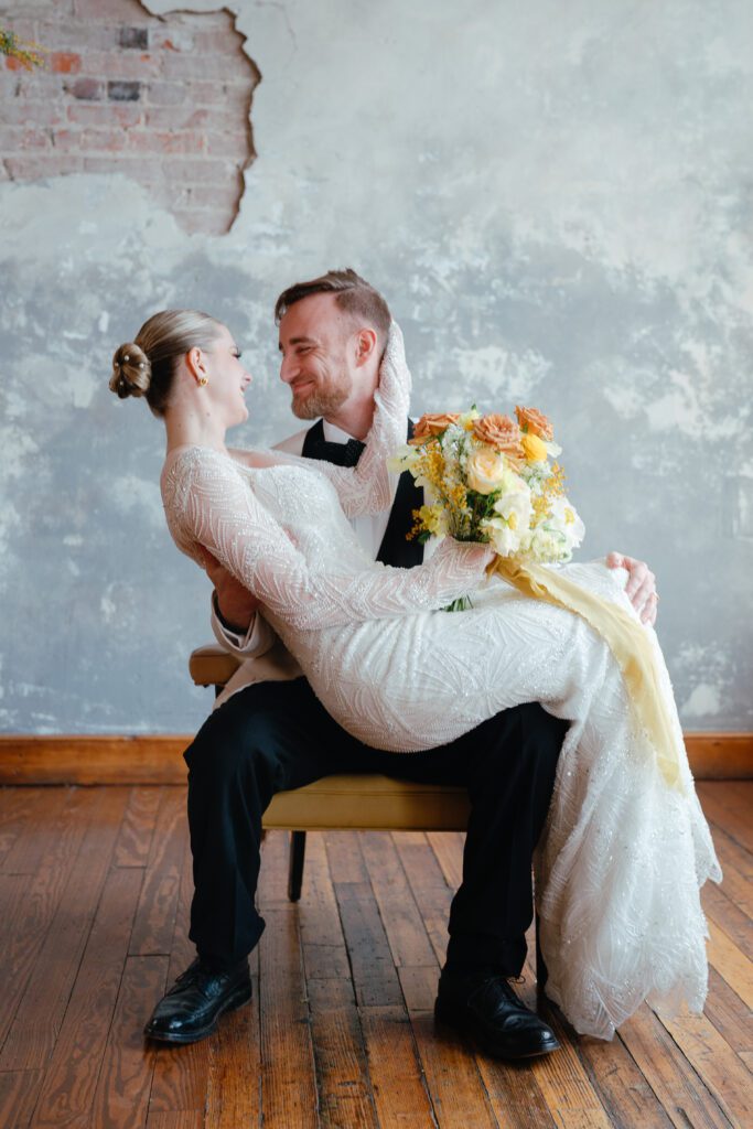 A woman in a wedding gown holding a bouquet of flowers sits on a grooms lap while they smile at one another.