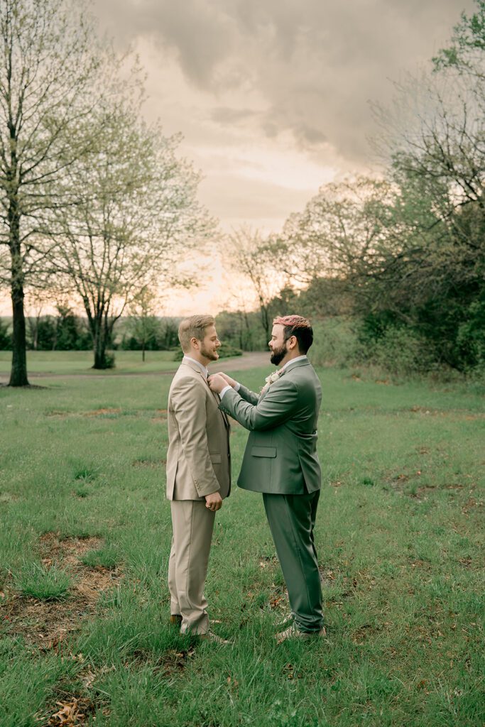 A groom in a green suit adjusts his new husbands tie while standing on the lawn of Deer Ridge Estates