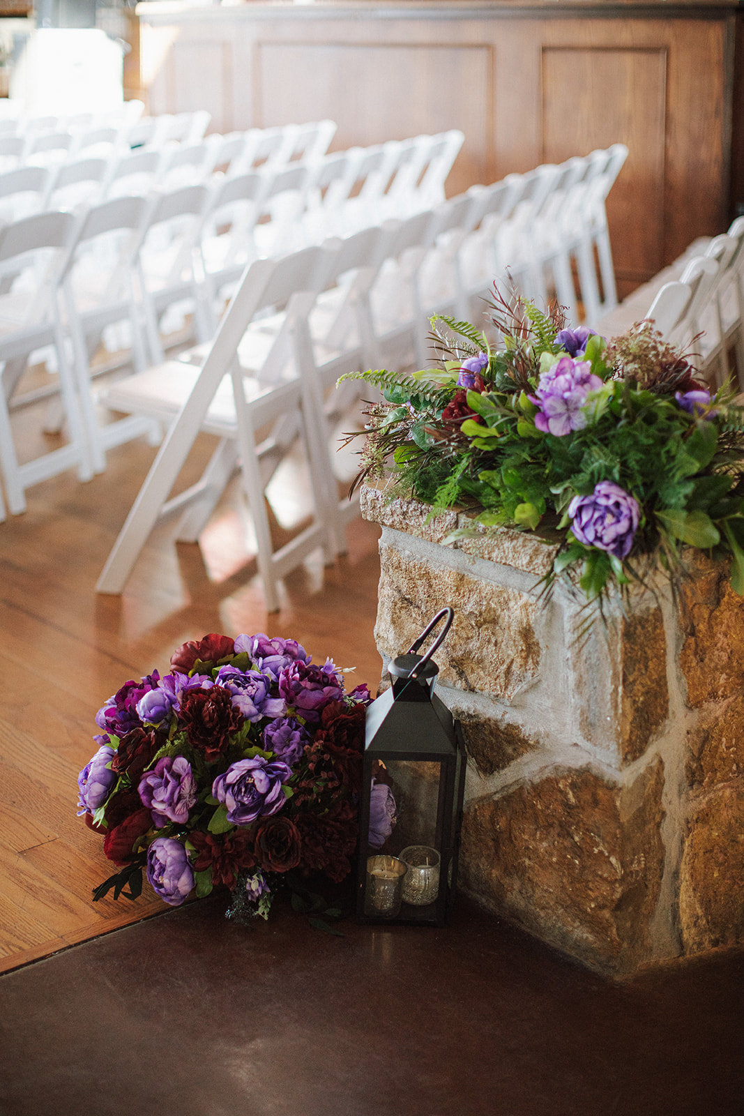 A floral arrangement sits on the floor next to a lantern. There is a stone column behind it with more florals on top. 